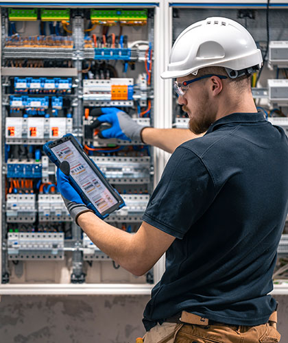 Electricista con casco, gafas y guantes revisando un cuadro eléctrico industrial con una tablet en la mano